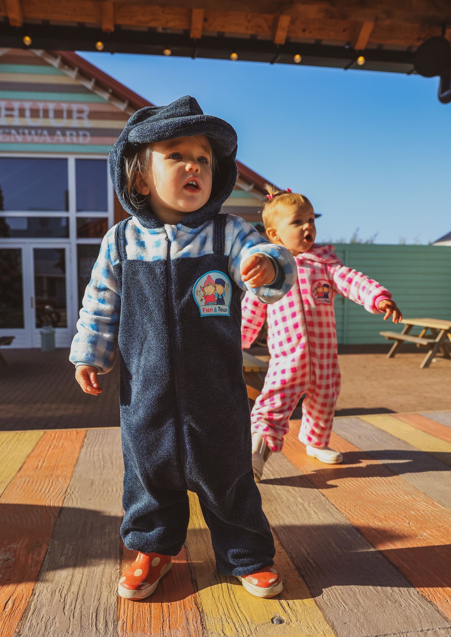 Two toddlers in overalls and playwear standing on wooden deck outside storefront. campagne shoot niuvwe collectie fotografie.