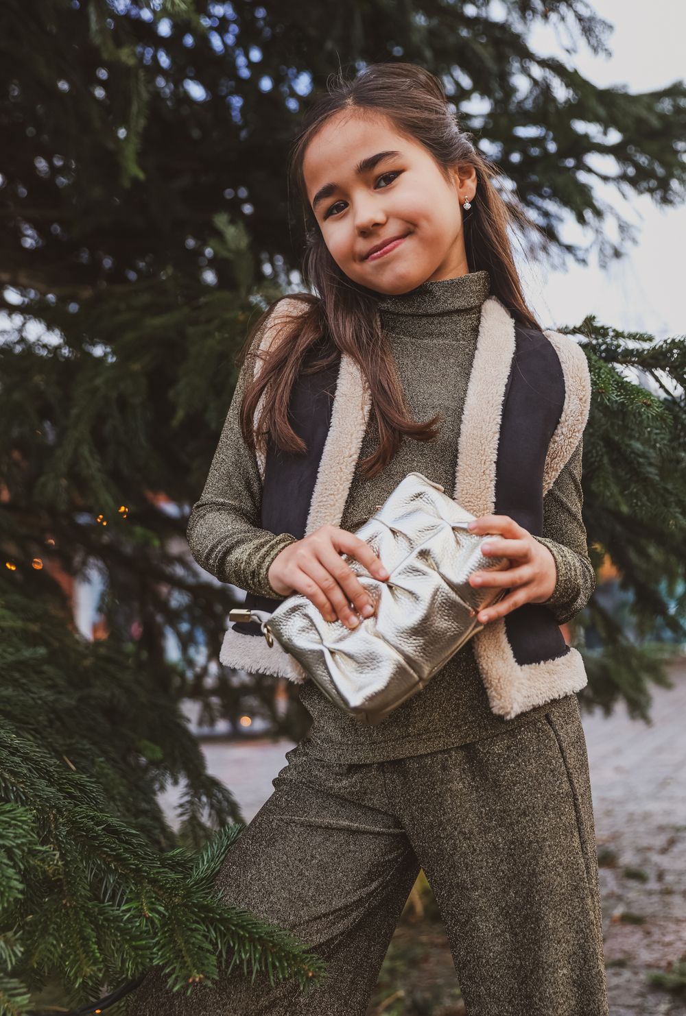Person in gray turtleneck holding silver gift while standing by evergreen tree.