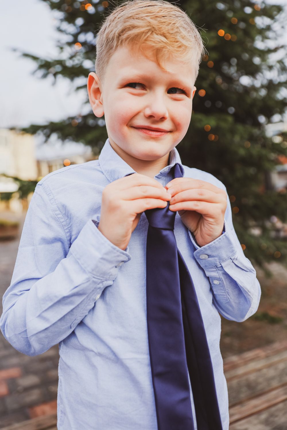 Young boy in light blue dress shirt adjusting navy tie outdoors.