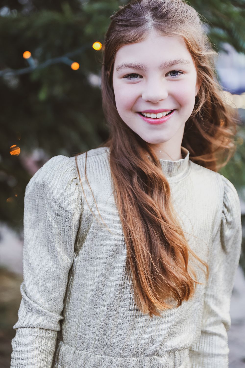 Person in cream sweater smiling with bokeh lights and evergreen branches in background.