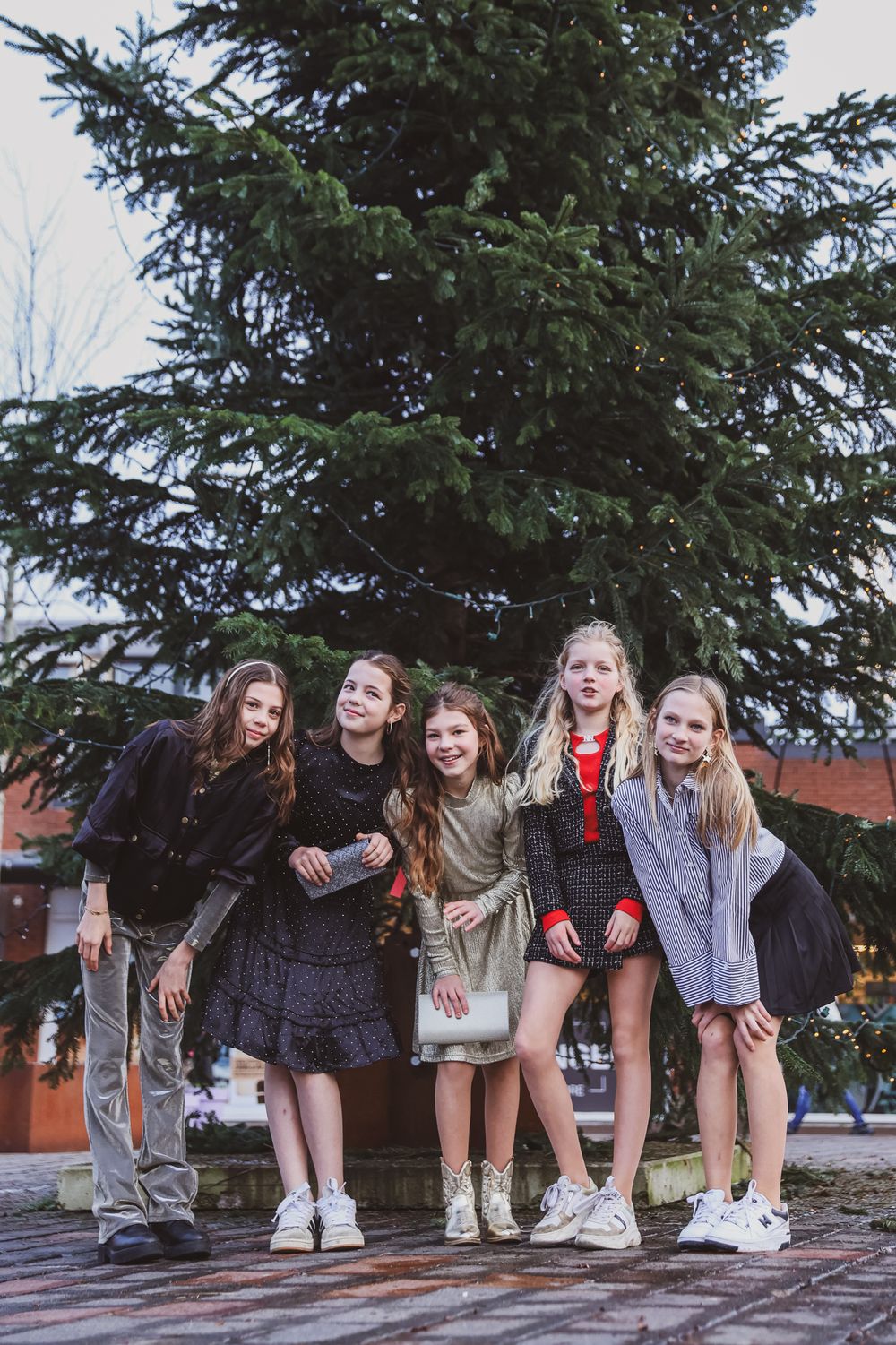 Five friends posing together outdoors in front of a large evergreen tree.