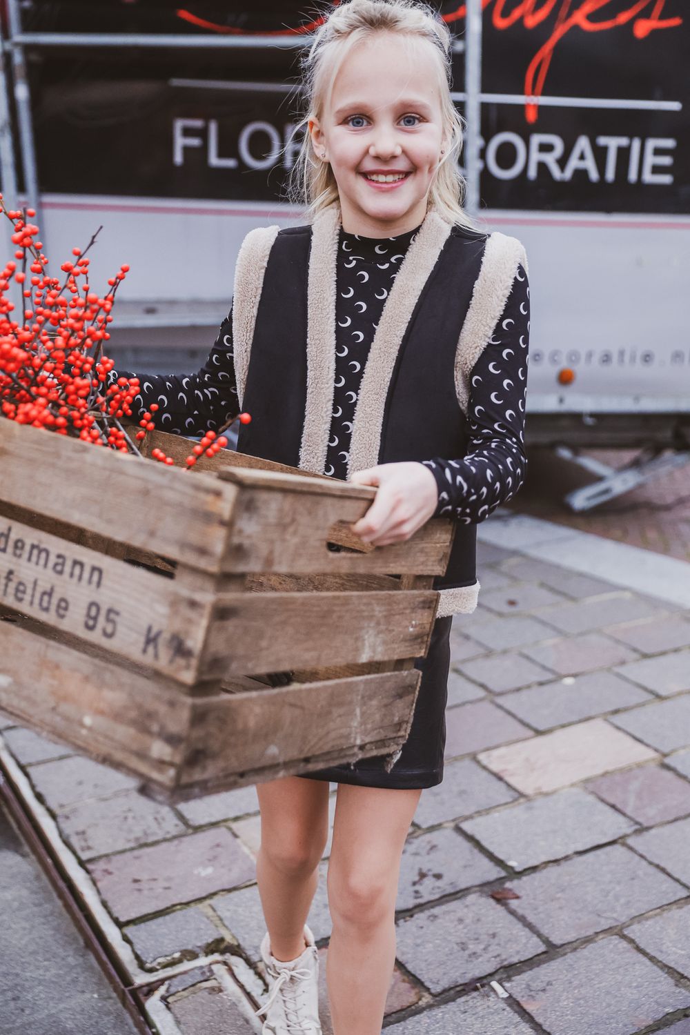 Young person carrying wooden crate with red berries near floral shop.
