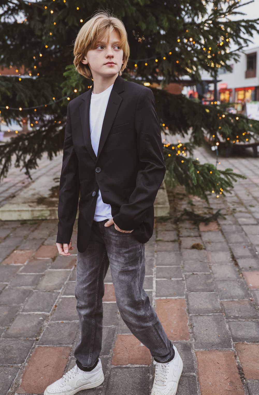 Young person in black blazer and jeans posing against Christmas tree backdrop.