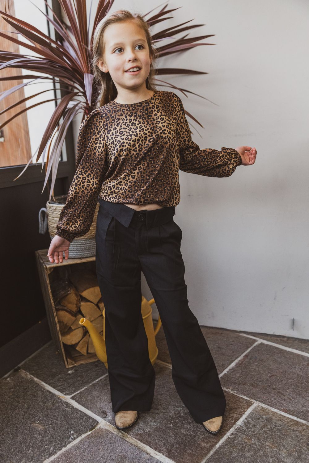 Person in leopard print top and black pants posing indoors near decorative plant.