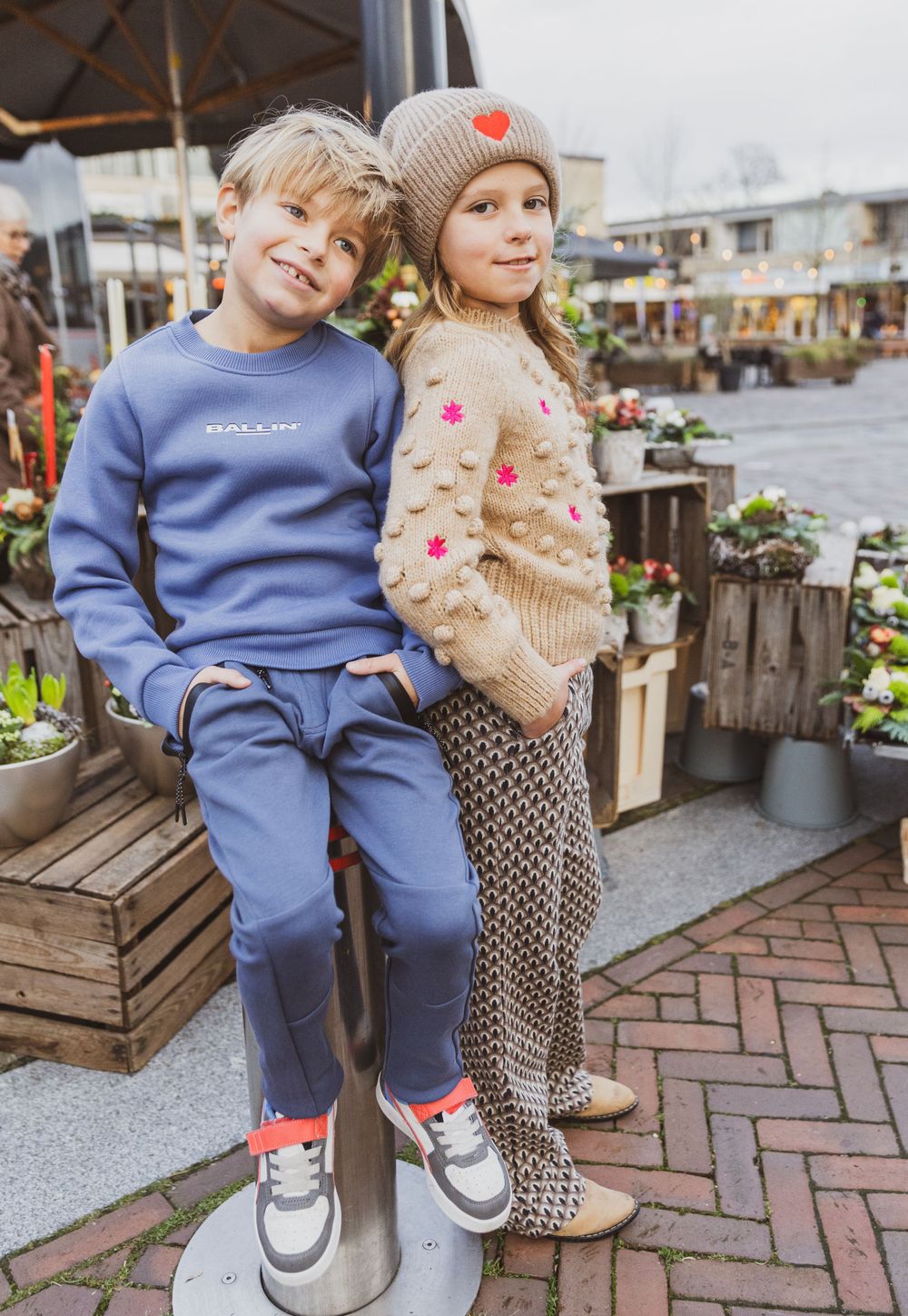 Two children in casual wear sitting together on wooden crates near outdoor garden displays.