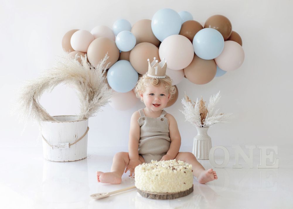 A happy toddler sits before a birthday cake with balloon arch and neutral decor in the background.