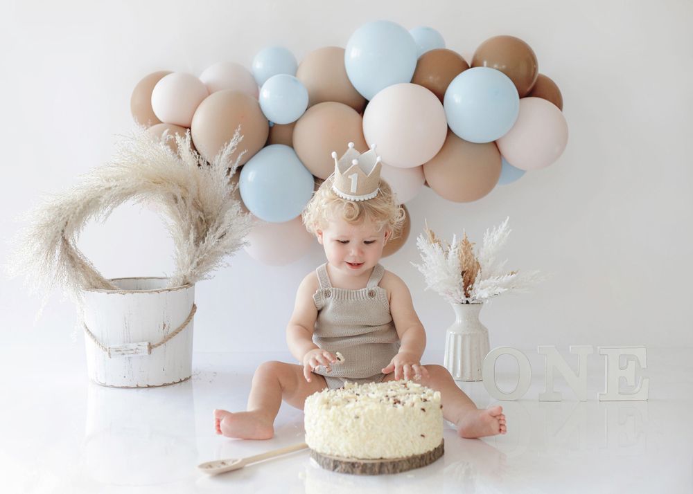 A delighted toddler sits with a white frosted cake against pastel balloon backdrop.