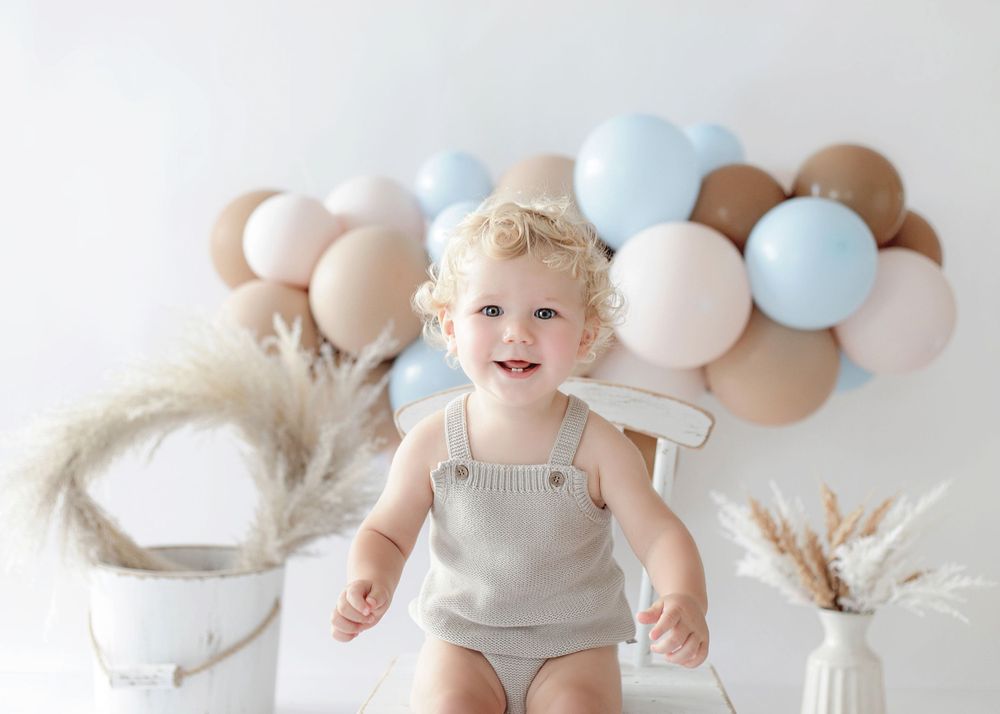 A small child in a beige romper poses against a pastel balloon backdrop with pampas grass decorations.