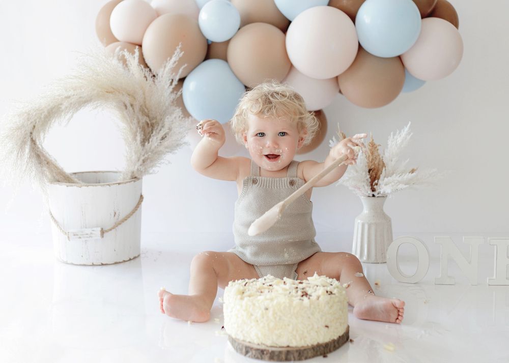 An excited toddler celebrates with arms raised near birthday cake with balloon arch backdrop.
