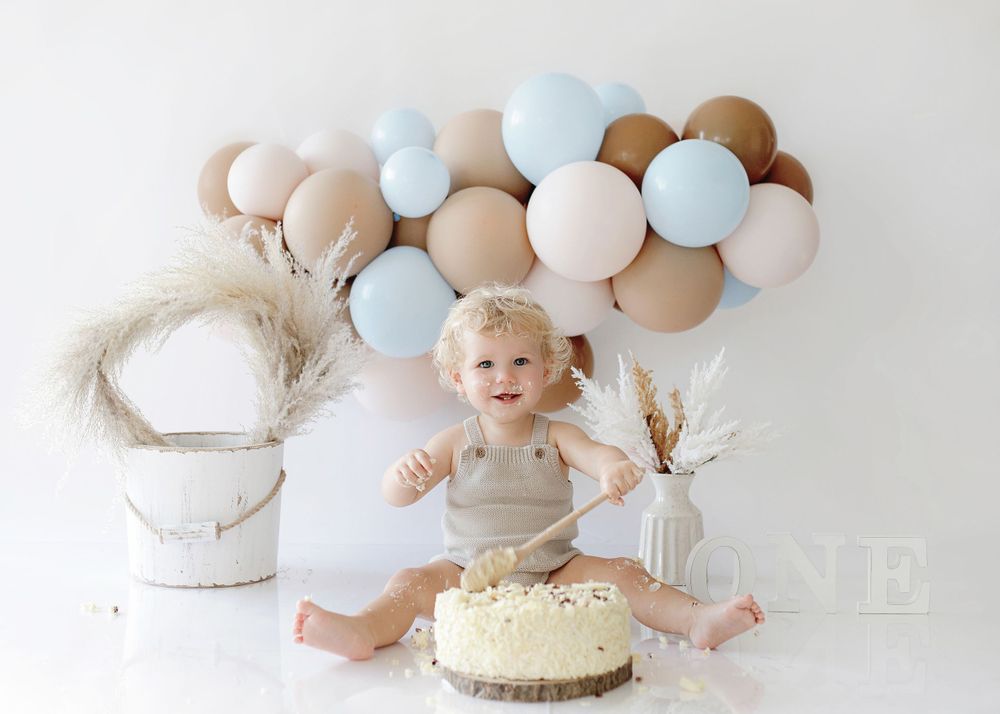 Baby in beige outfit sits excitedly in front of birthday cake with light blue and beige balloon arch decoration.