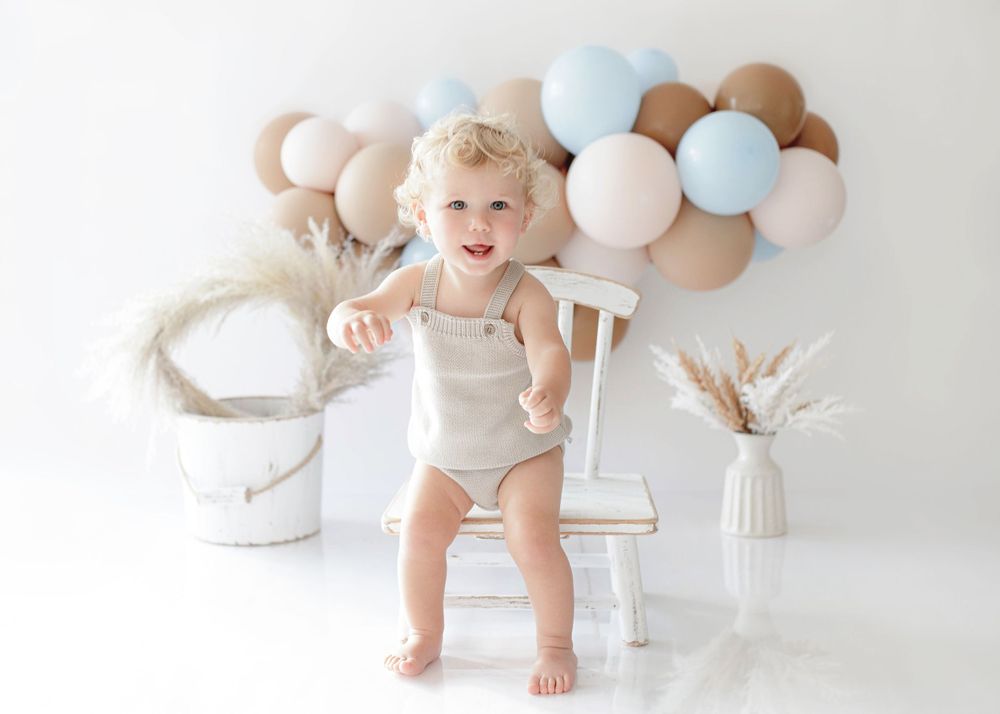 A joyful baby stands on a white chair with soft balloons and pampas grass decor in the background.