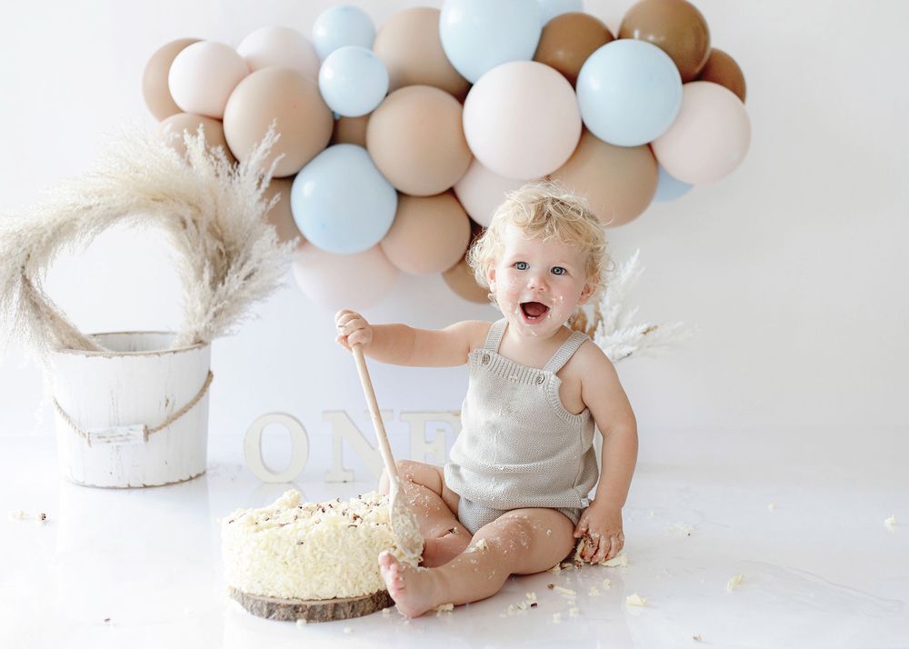 Baby smiling and reaching toward birthday cake decorated with light blue and beige balloons in background.
