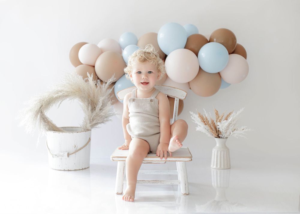 A toddler sits on a white chair against a wall decorated with tan and blue balloons and dried flowers.