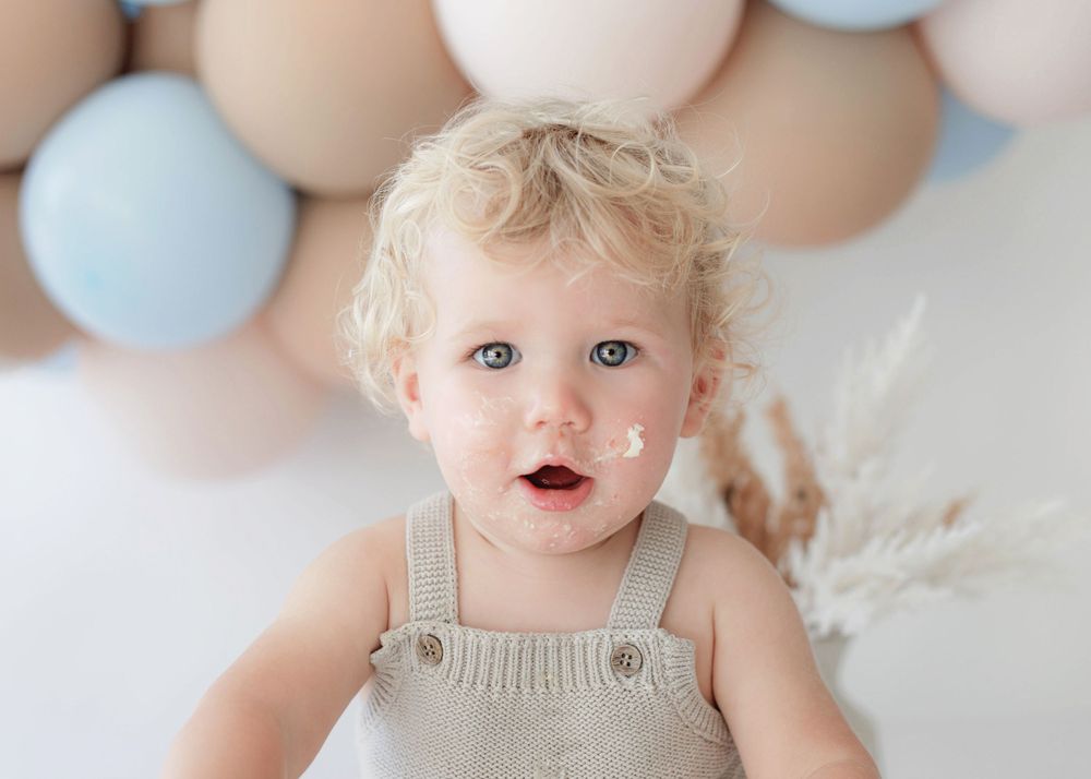 Child wearing beige overalls sits in front of soft blue and tan balloon arch backdrop.
