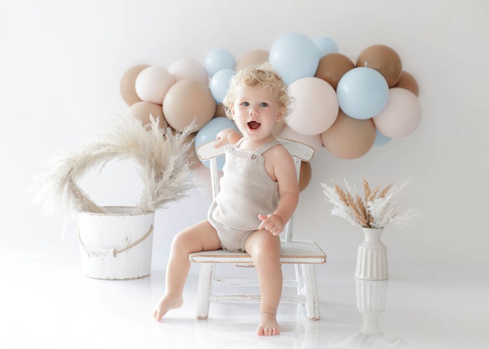 A happy toddler in gray romper sits on white chair with balloon arch and pampas grass decorations.