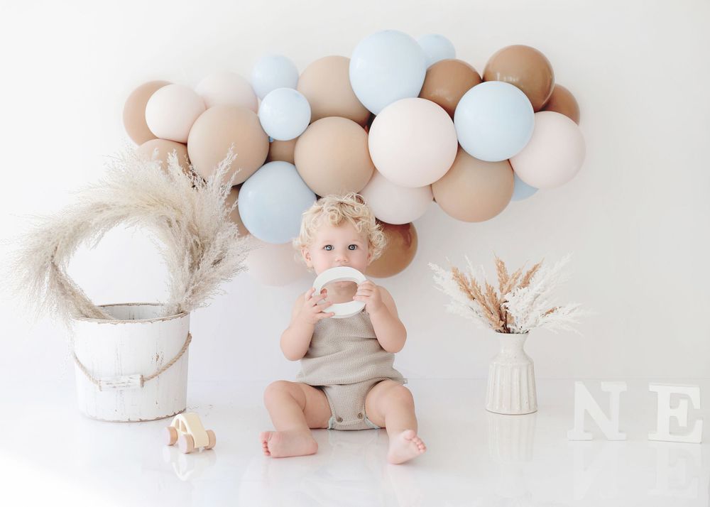 A happy baby in beige outfit sits with balloons and decorative elements in a neutral toned photoshoot.