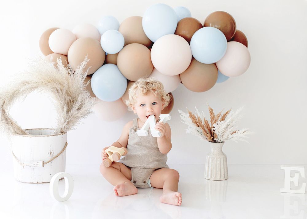 A smiling toddler in beige romper poses with pastel balloon arch and dried floral decorations.