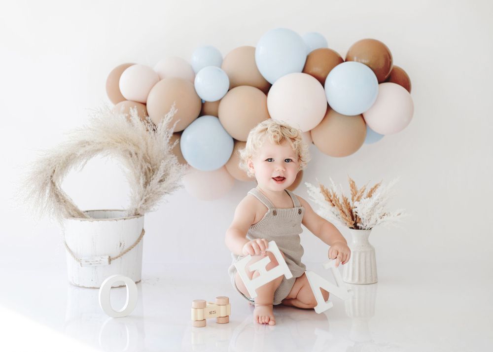 A playful toddler sits among wooden blocks with balloon garland and pampas grass decorations.