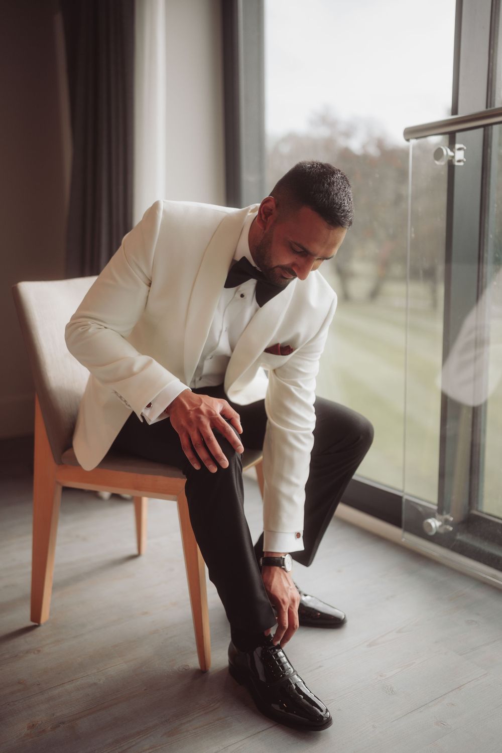 A person in a white tuxedo jacket and black pants sits thoughtfully on a chair near a window.