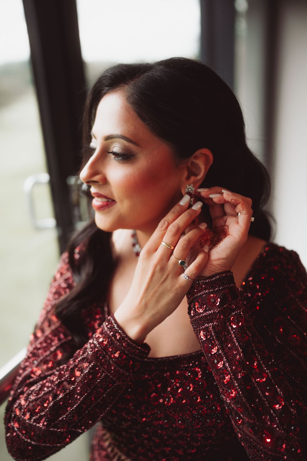 Woman in red sequined dress adjusts her earring while posing near a window.