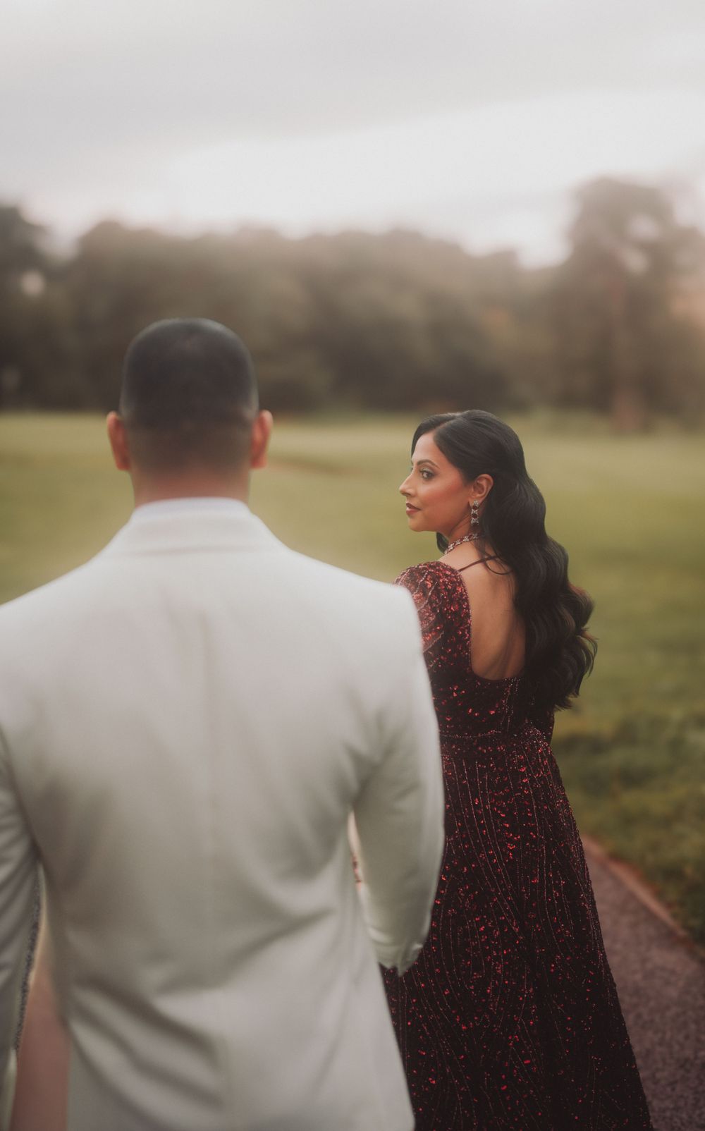 A couple in formal attire poses in a misty outdoor garden setting with the woman in a sparkling burgundy gown.