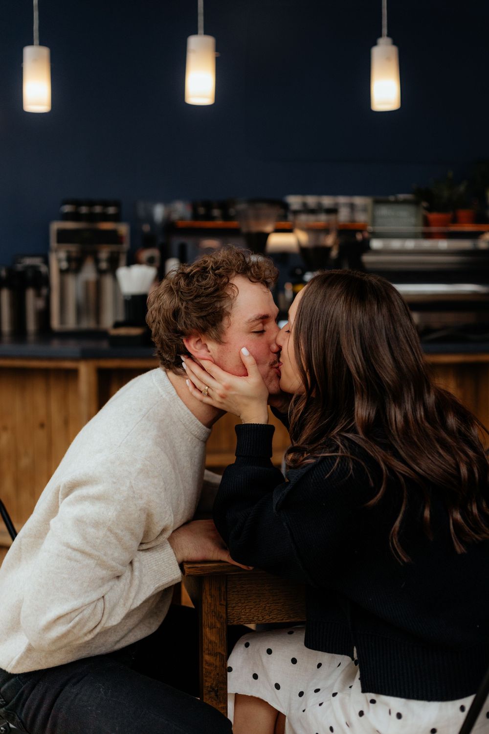 A romantic couple shares a tender kiss while sitting at a coffee shop counter with pendant lights hanging above.