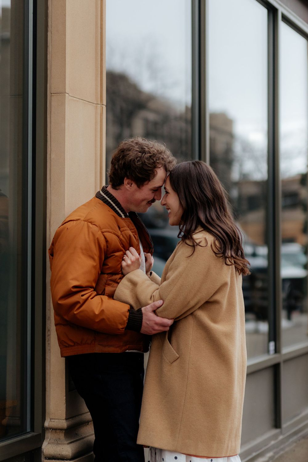 A romantic couple wearing winter coats shares an intimate moment together while standing against a building exterior.