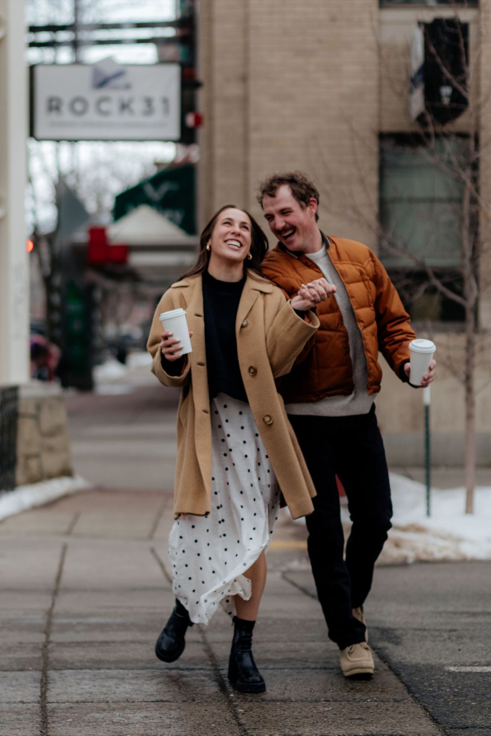Two people walking down a city street laughing and holding coffee cups on a winter day.