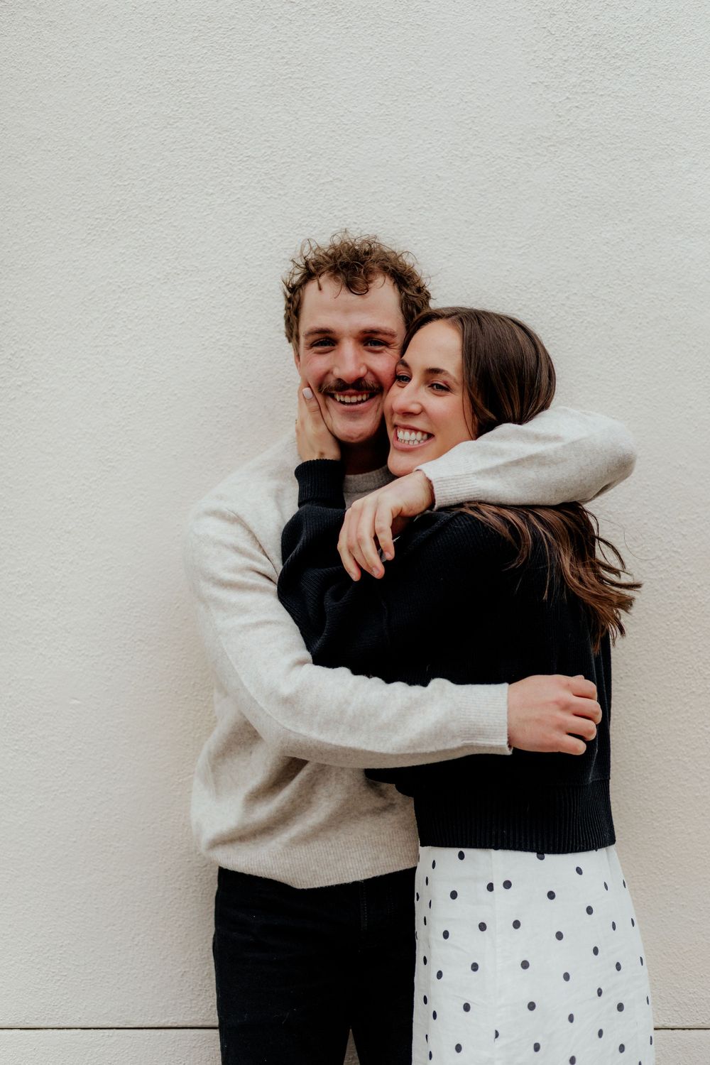 A couple embraces and laughs while standing against a white wall, wearing casual sweaters and a polka dot skirt.