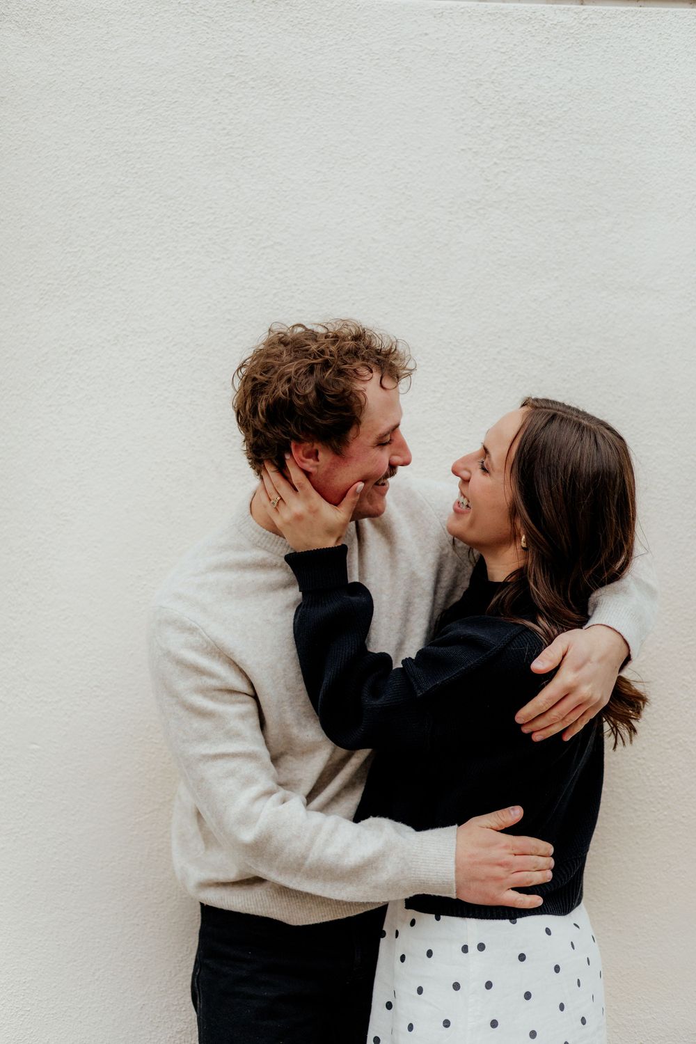 A romantic moment between a couple wearing black and cream colored outfits against a white wall.