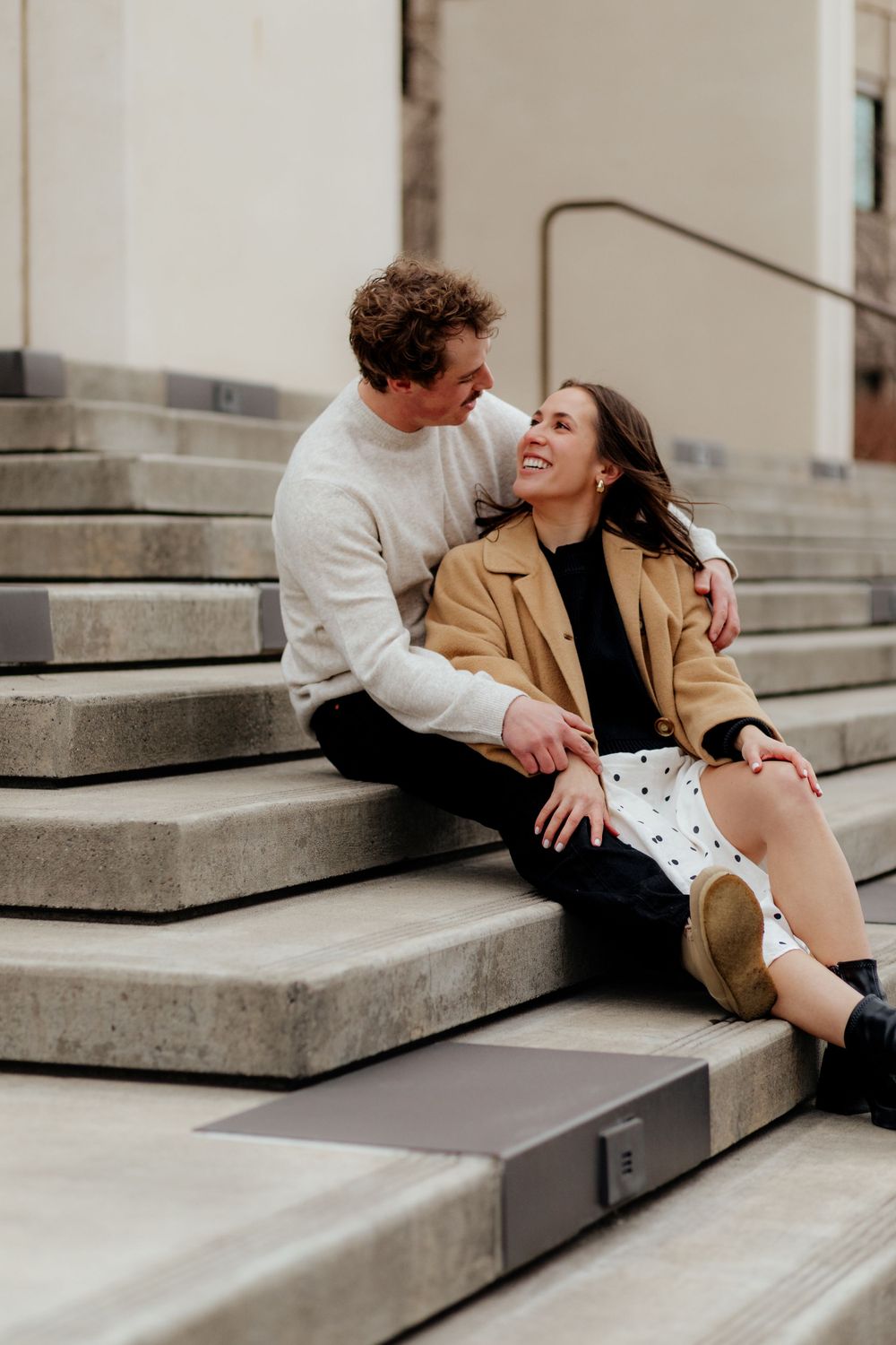 A couple sits closely together on concrete steps, sharing an intimate moment in casual winter attire.