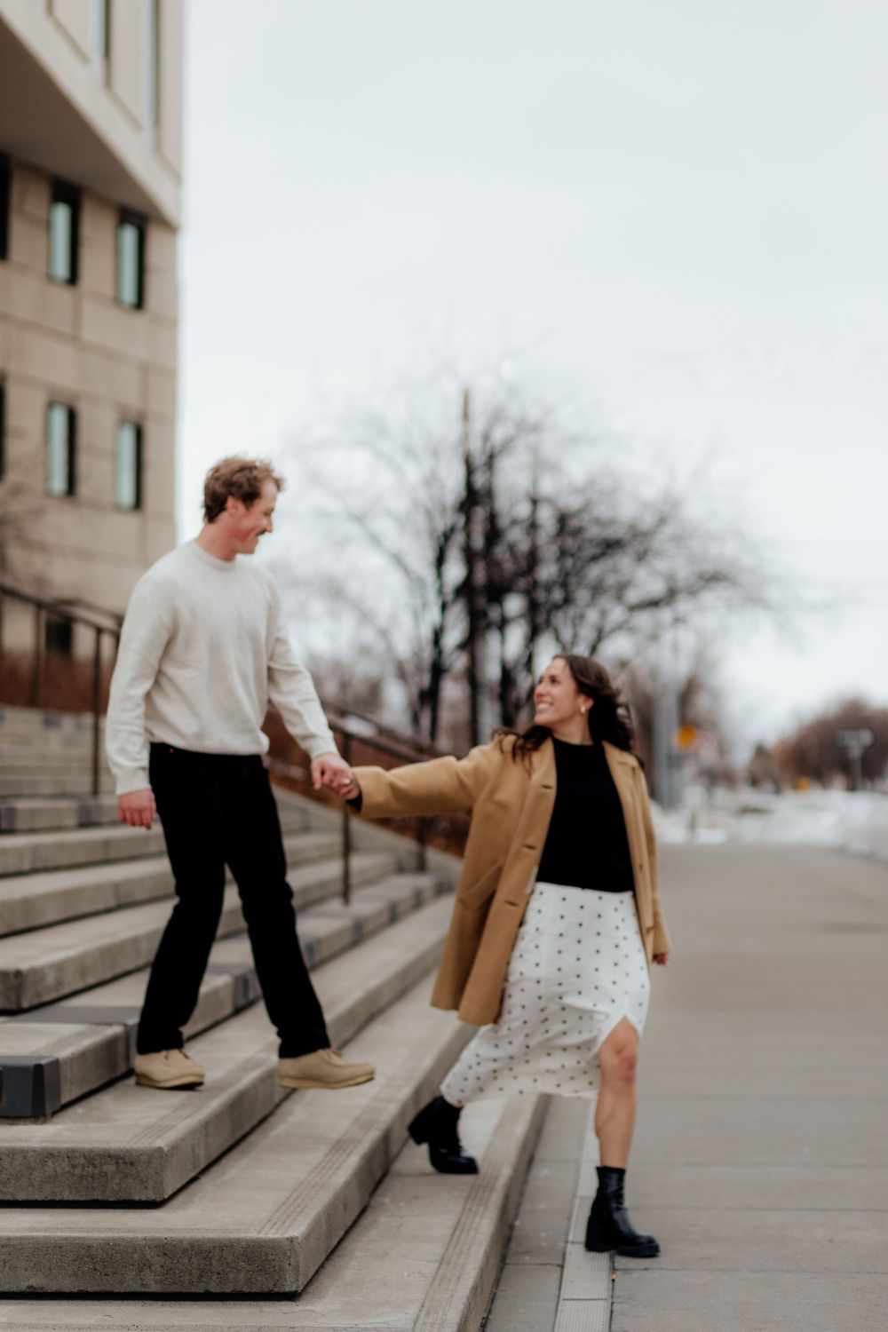 A couple shares a playful moment on concrete steps with one person in a camel coat and another in a cream sweater.