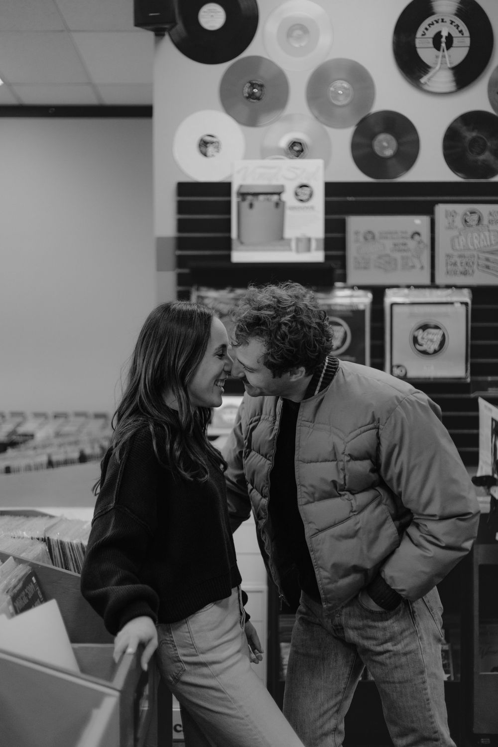 A couple shares an intimate moment while browsing vinyl records in a music store with records displayed on the wall.