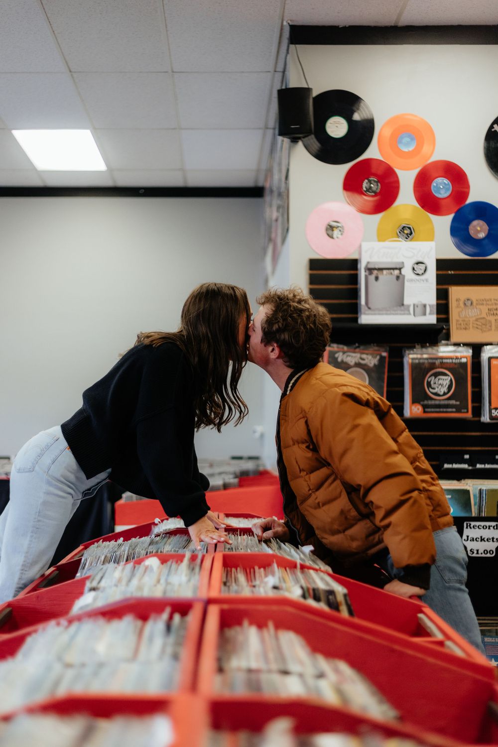 Two people browse through vinyl records at a music store with colorful vinyl discs decorating the wall.