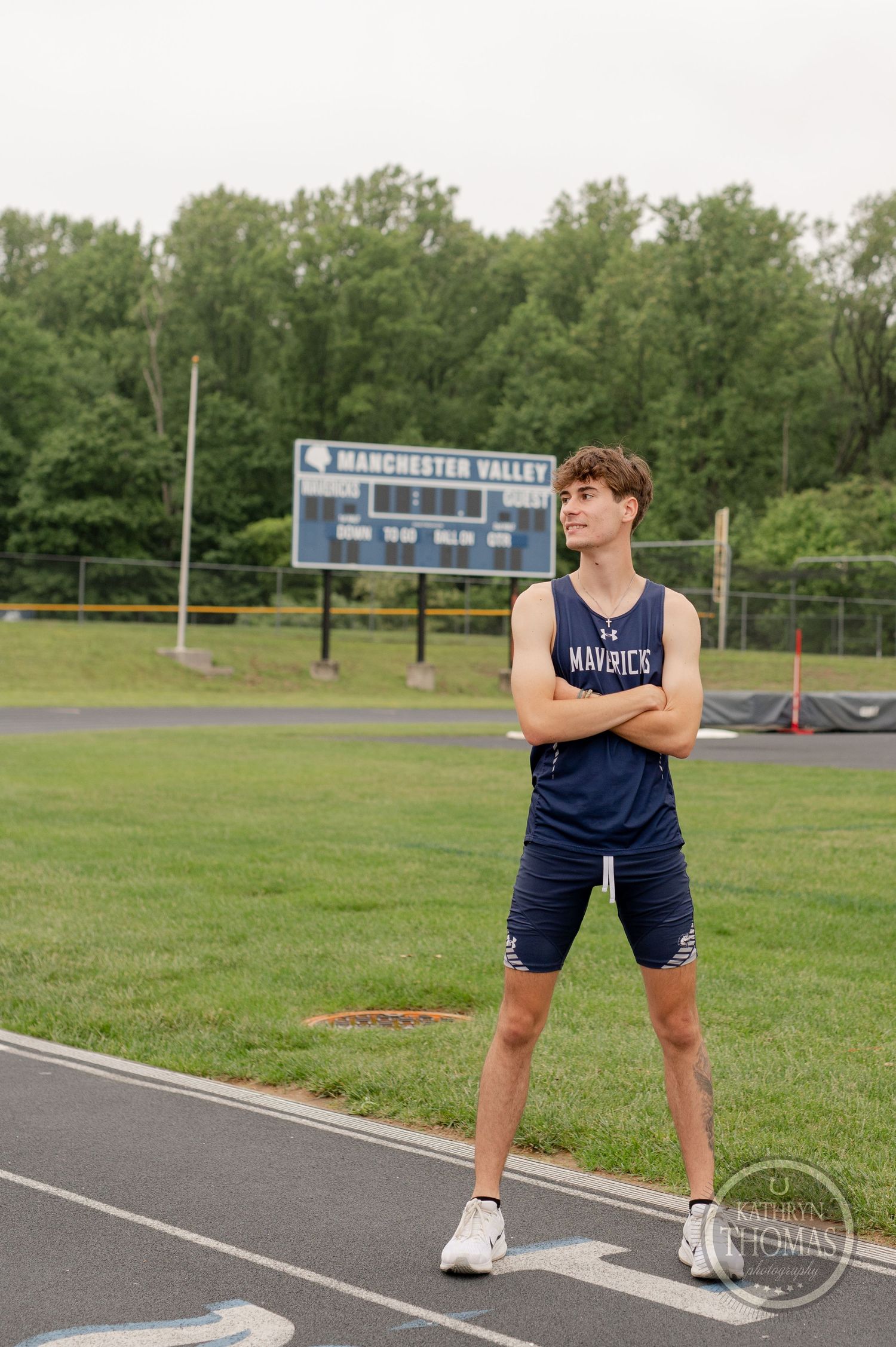 Track athlete in navy uniform stands confidently on running track near scoreboard.