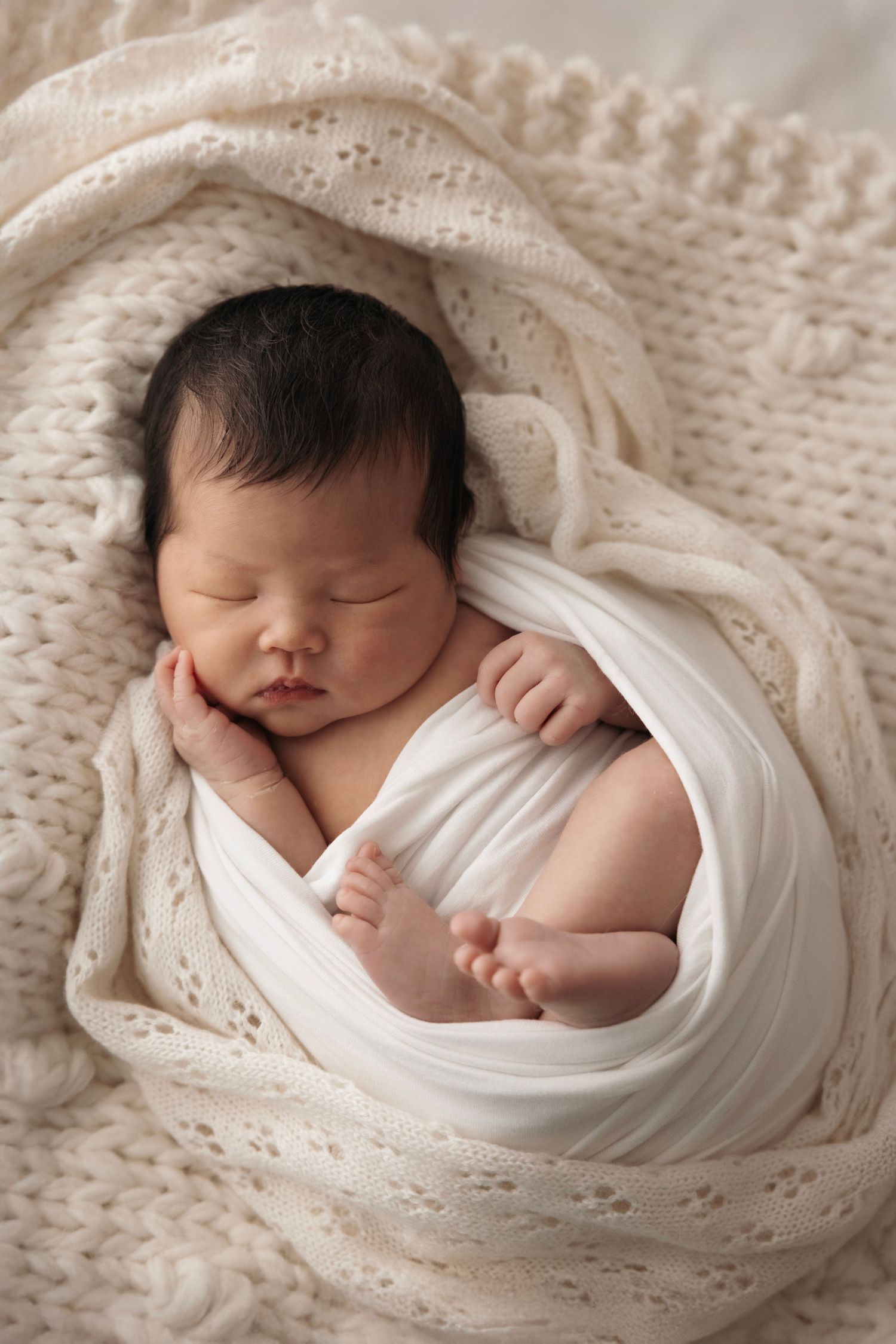 A sleeping newborn baby wrapped in white fabric lies on a cream-colored knit blanket in a professional photography setting.