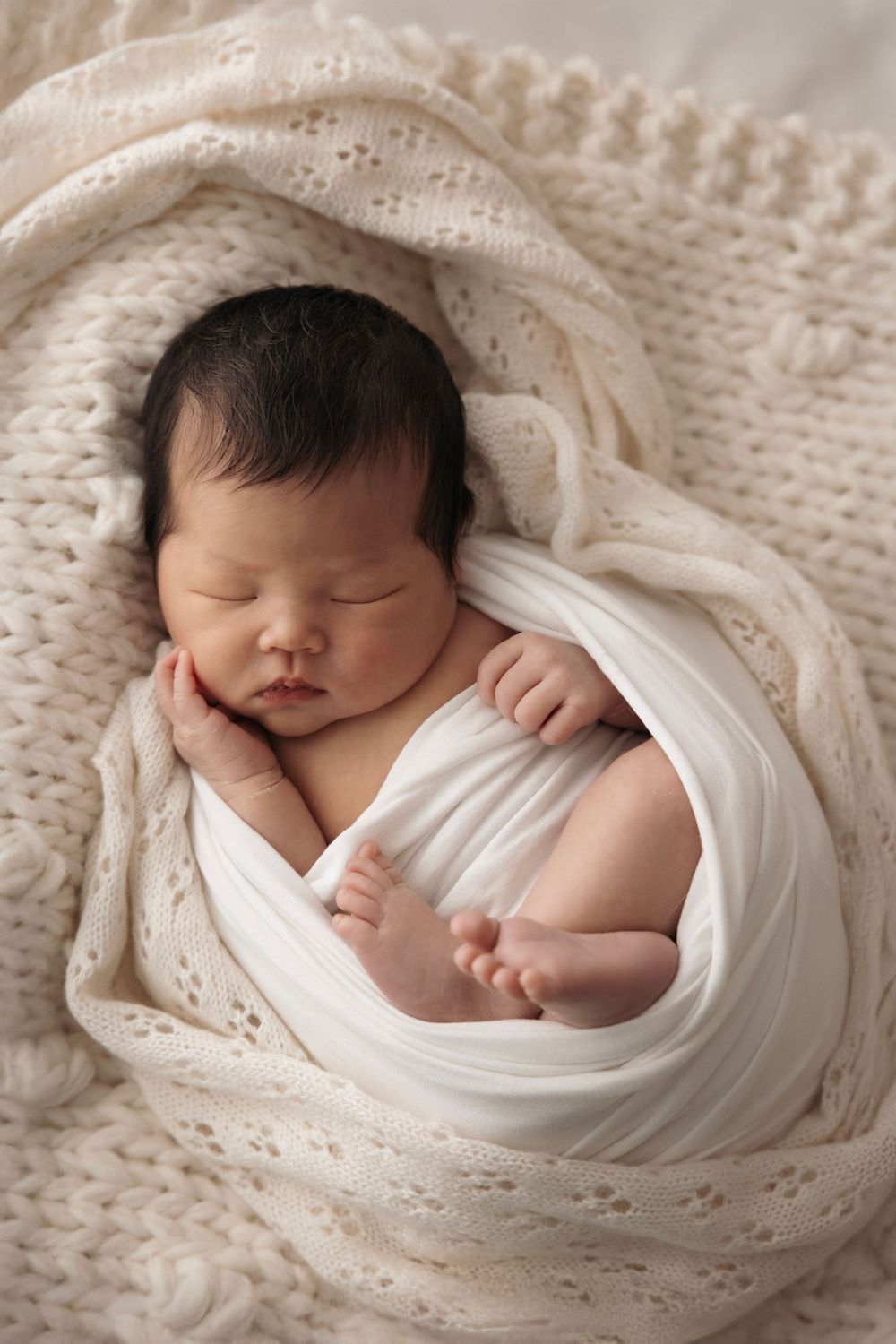 A sleeping newborn baby wrapped in white fabric lies on a cream-colored knit blanket in a professional photography setting.