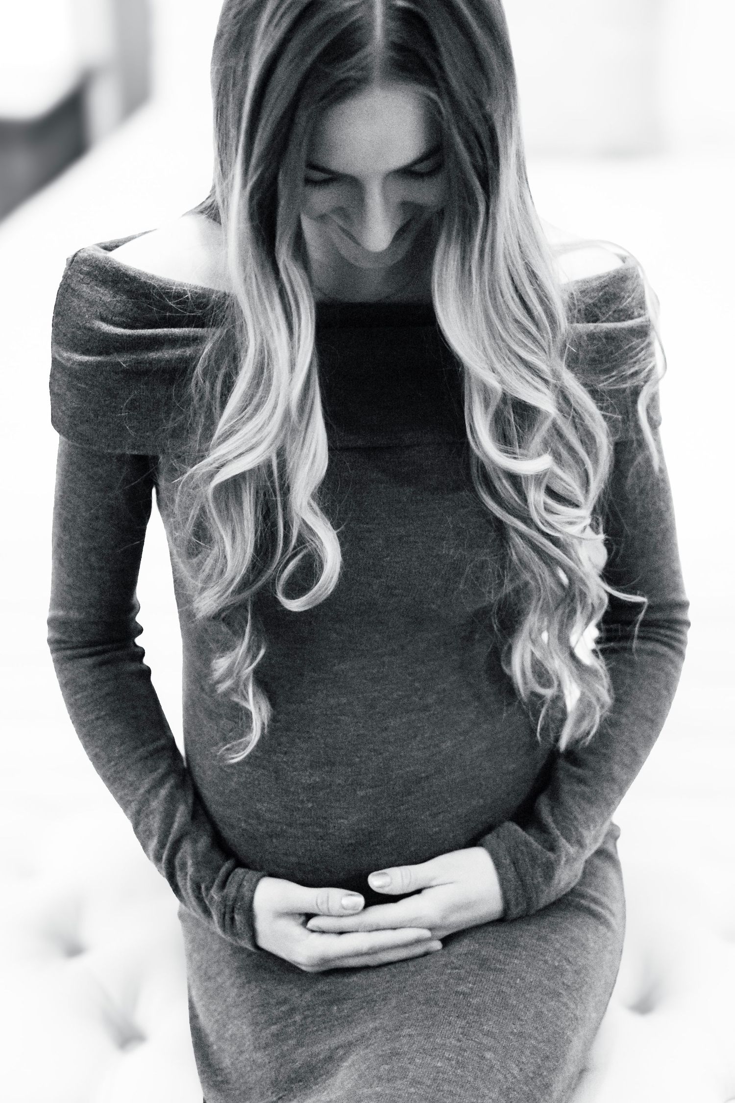 Black and white portrait of someone with long wavy blonde hair wearing a dark dress while cradling their stomach.