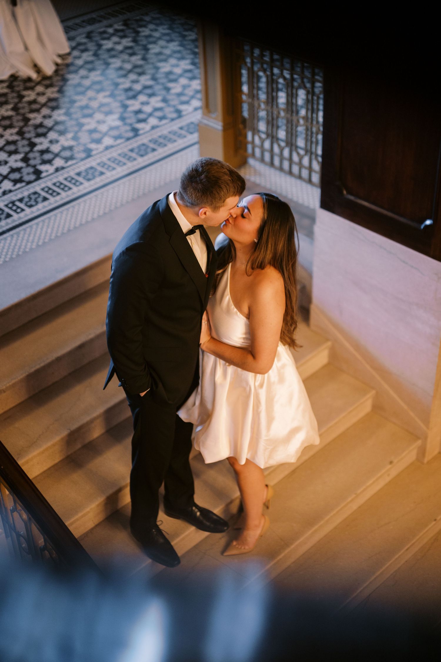 Couple sharing a romantic kiss on elegant marble stairs with ornate patterned floor and window in the background.