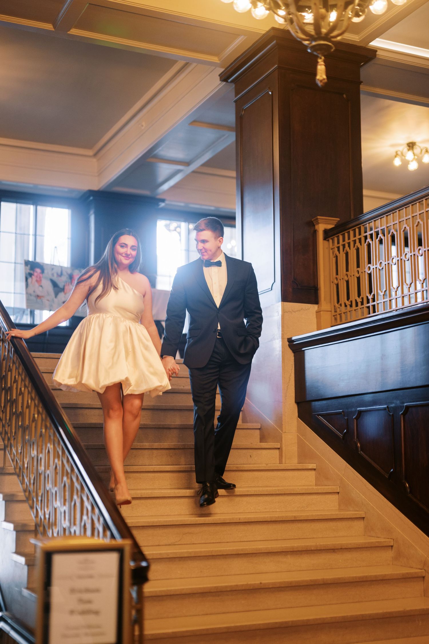 A couple descends an elegant wooden staircase, with the woman wearing a cream dress and man in dark suit and yellow tie.