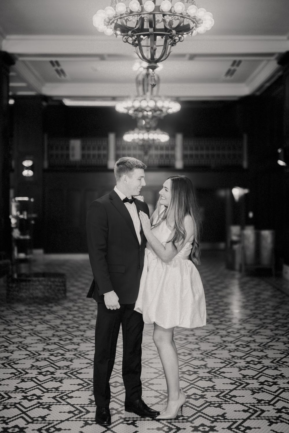 A couple in formal attire sharing an intimate moment under ornate crystal chandeliers on a patterned ballroom floor.