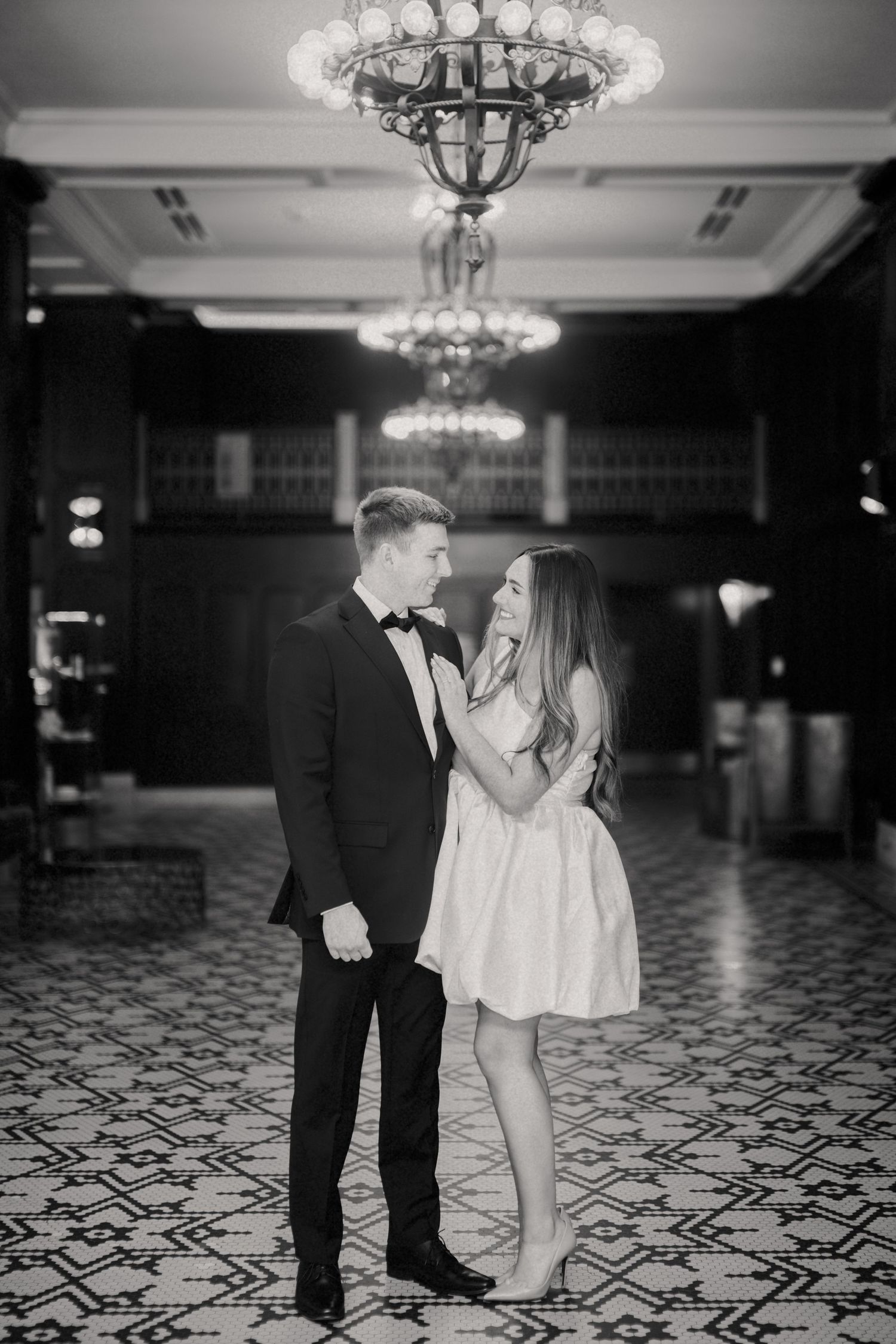 A couple in formal attire sharing an intimate moment under ornate crystal chandeliers on a patterned ballroom floor.
