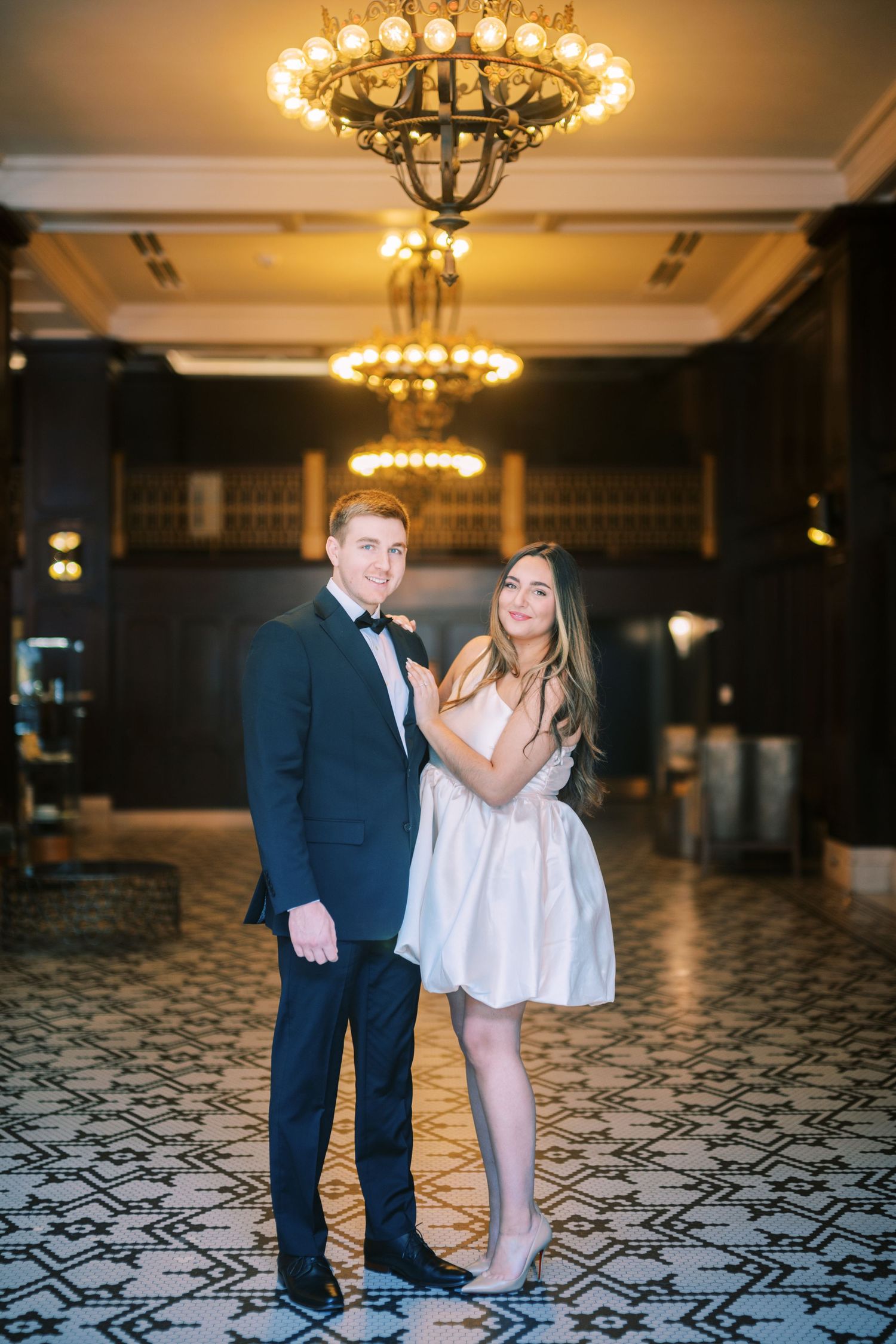 A couple poses together in formal attire under elegant chandeliers at an upscale venue with patterned floor.