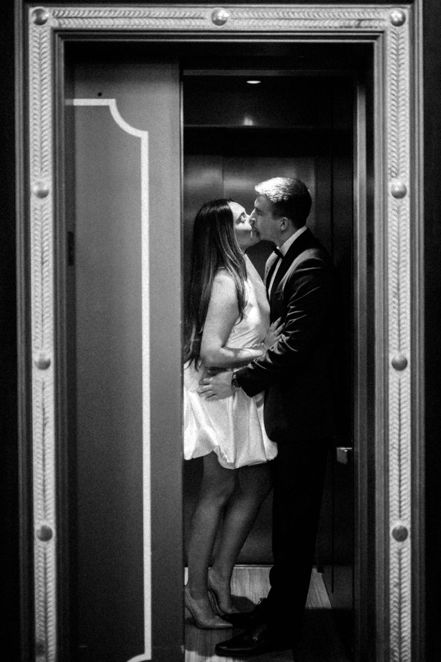 A romantic black and white photograph of a couple sharing an intimate kiss in an elevator doorway.