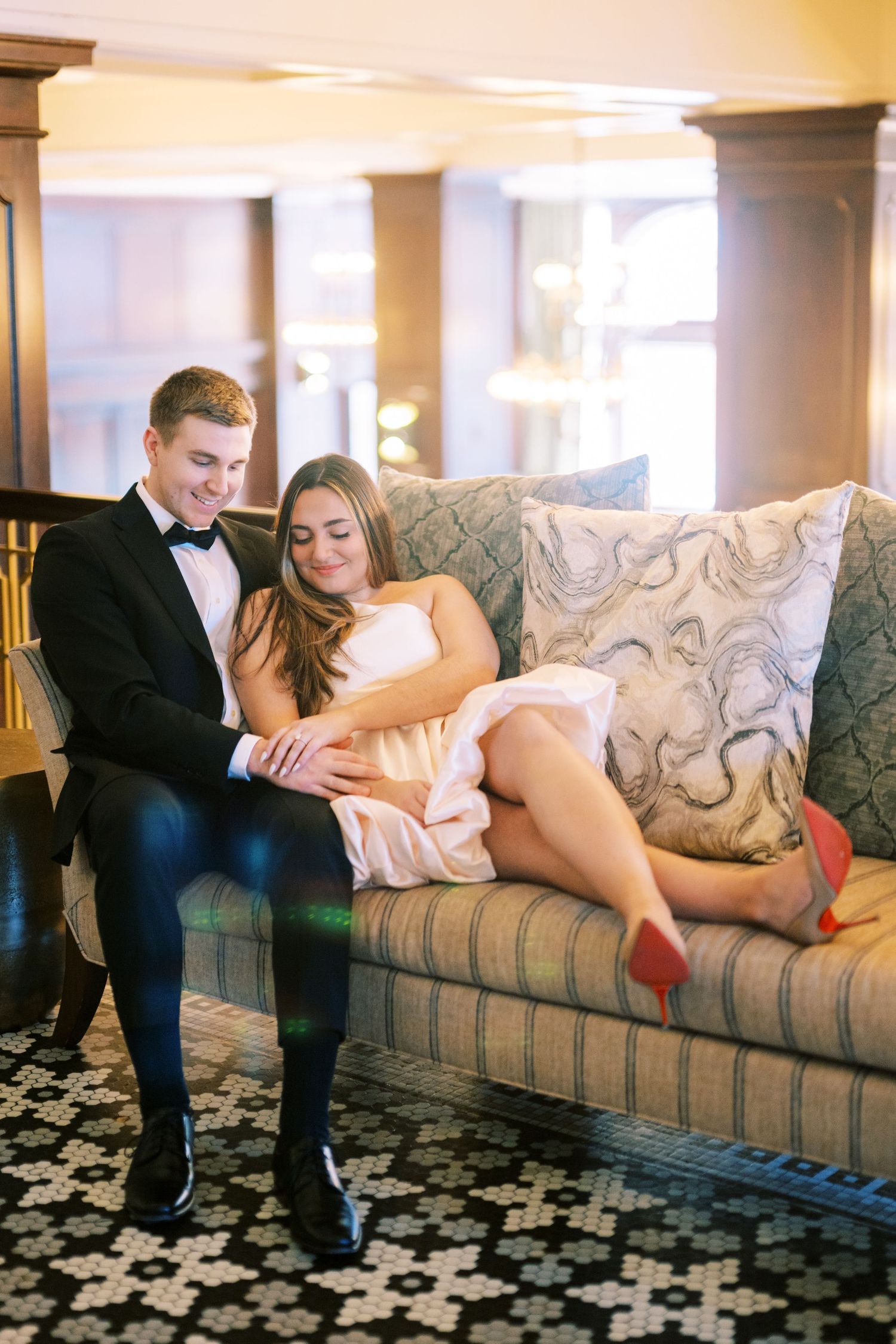 A couple in formal attire relax together on an elegant patterned couch in a warmly lit room.