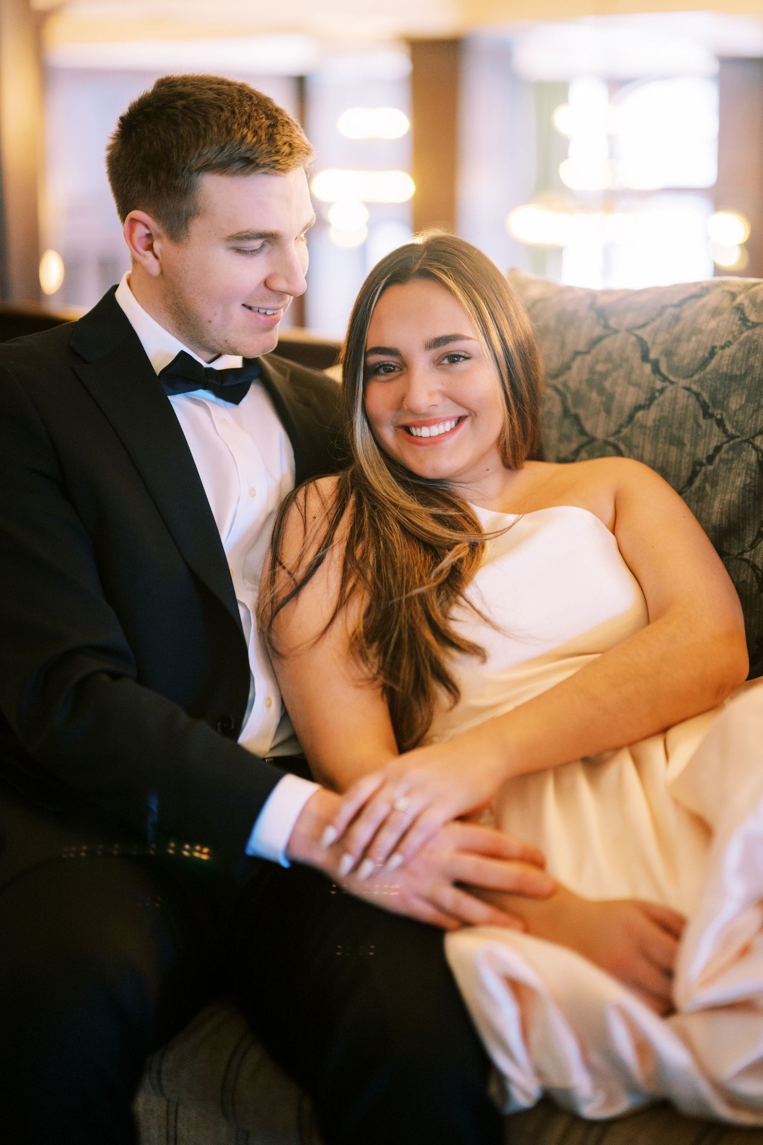 A couple in formal attire smiles while sitting close together on an elegant patterned couch.