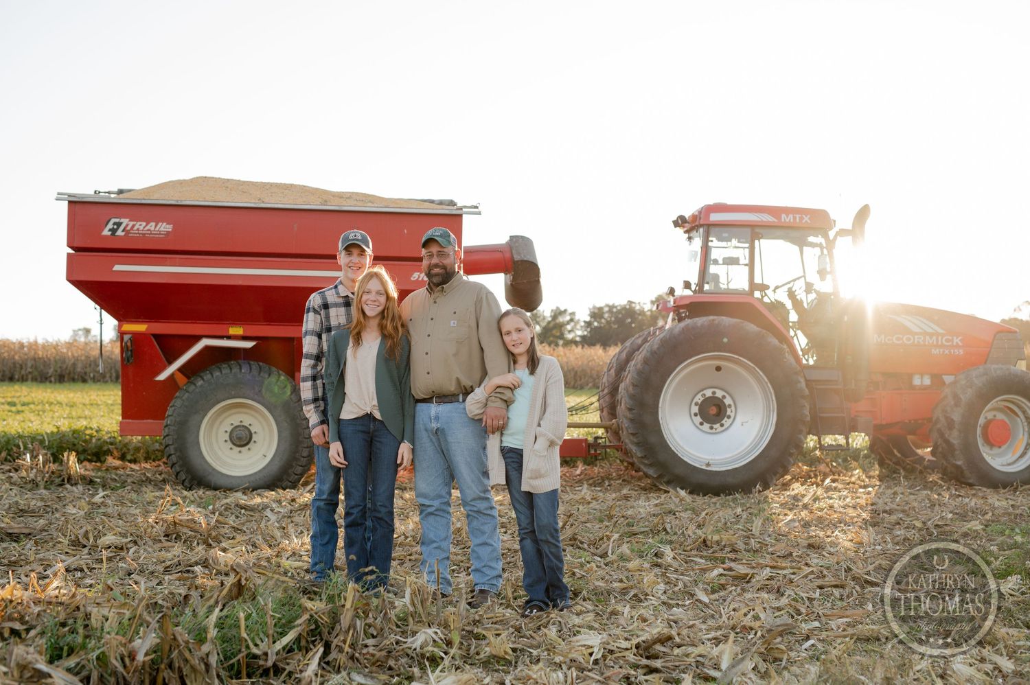 Farm workers pose next to a red grain cart and tractor during sunset on agricultural land.
