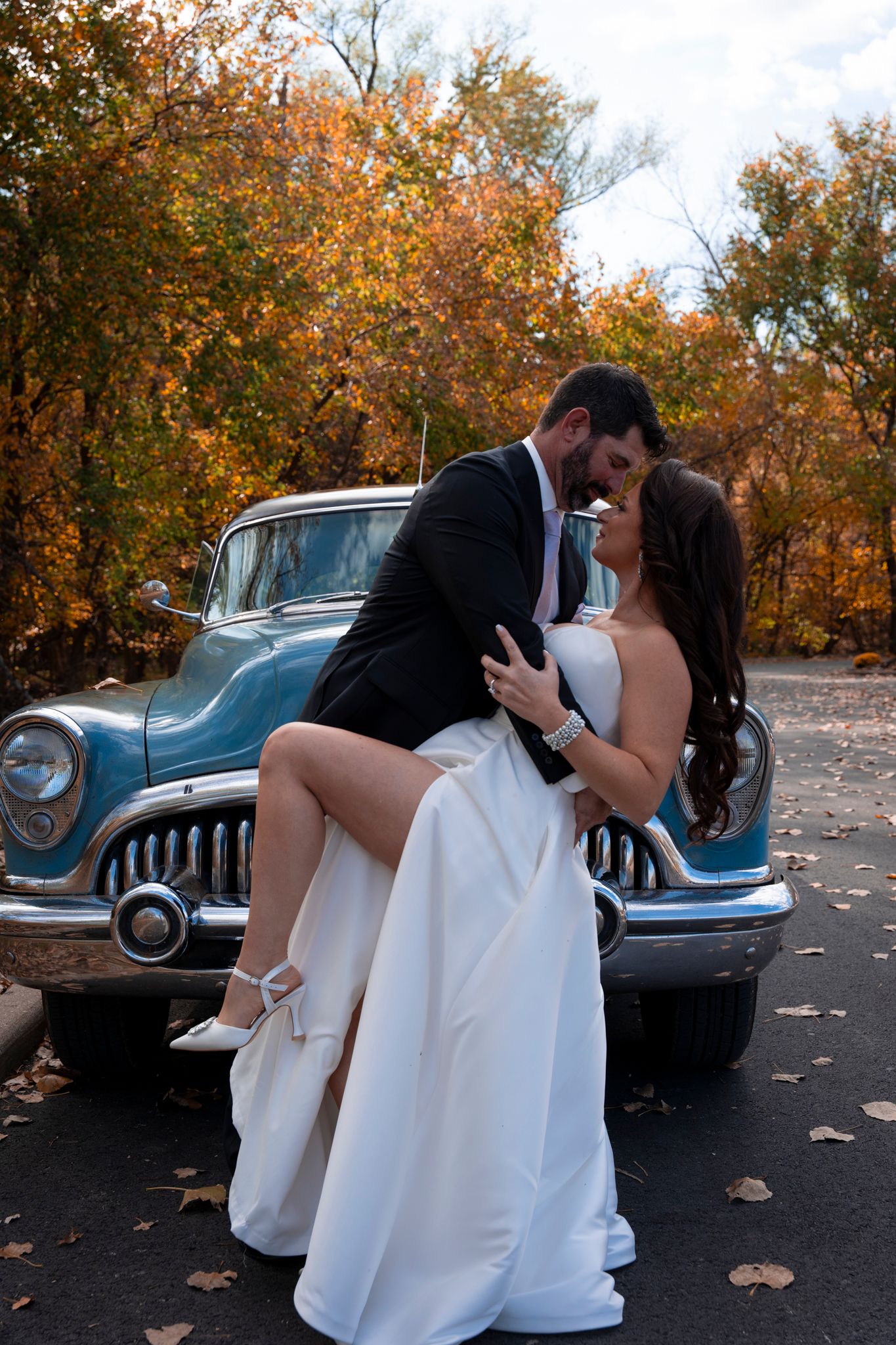 A romantic wedding portrait in front of a vintage blue car surrounded by autumn foliage.