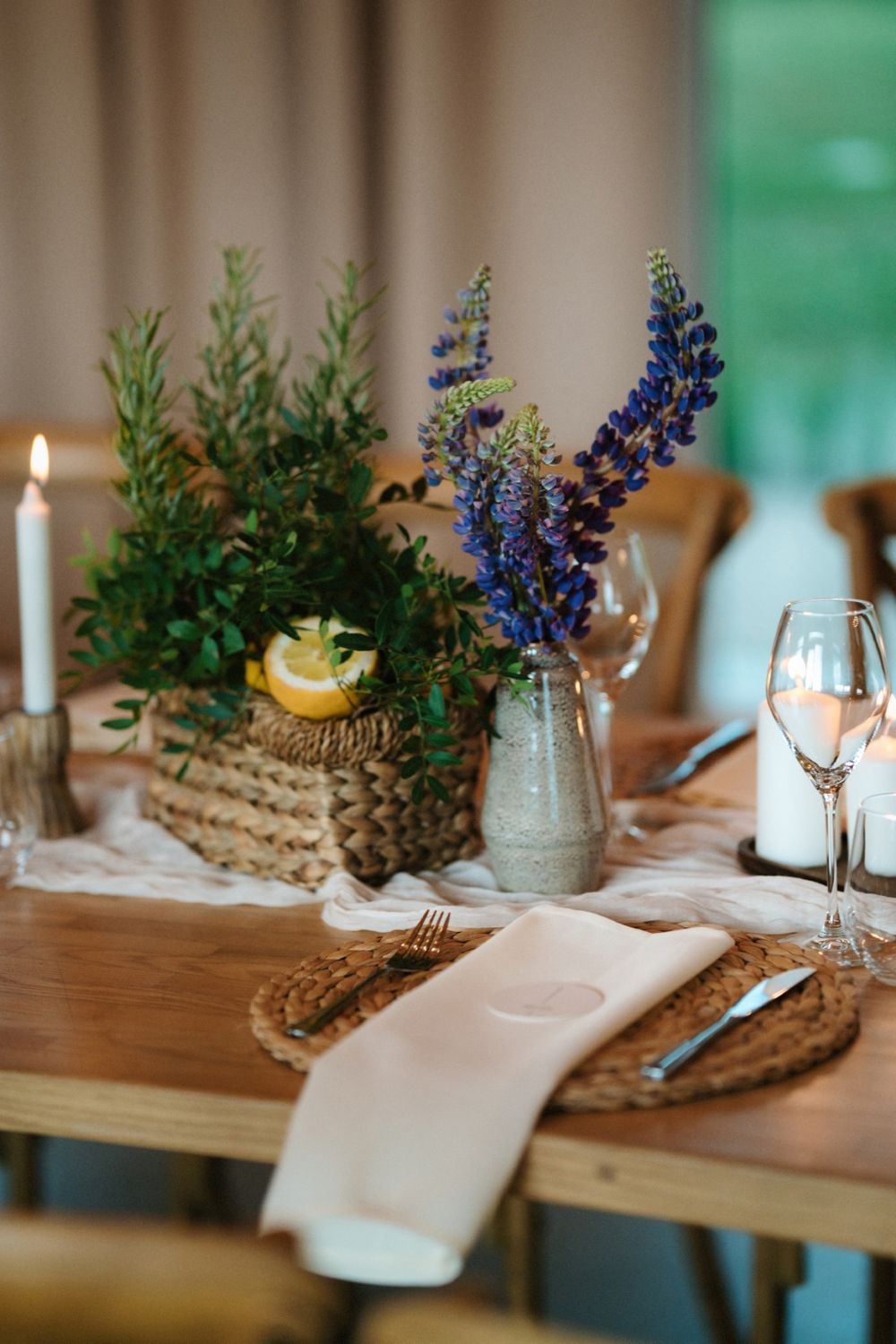 Elegant table setting with blue delphiniums in a vase, woven placemats, and white candles on a wooden surface.