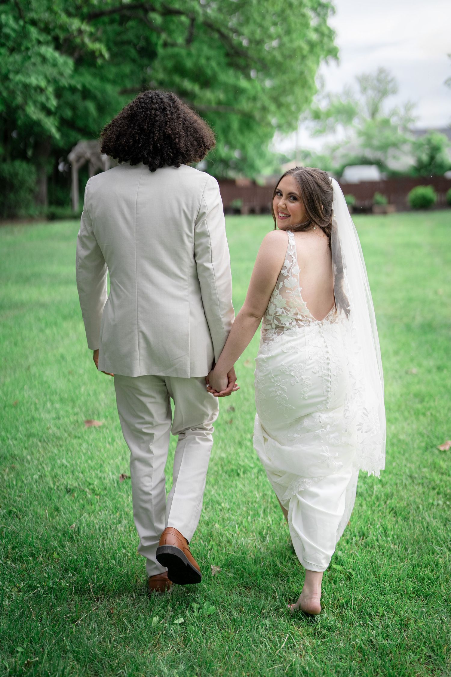 A wedding couple walks hand in hand across a lush green lawn, showing only their lower halves in a romantic garden setting.